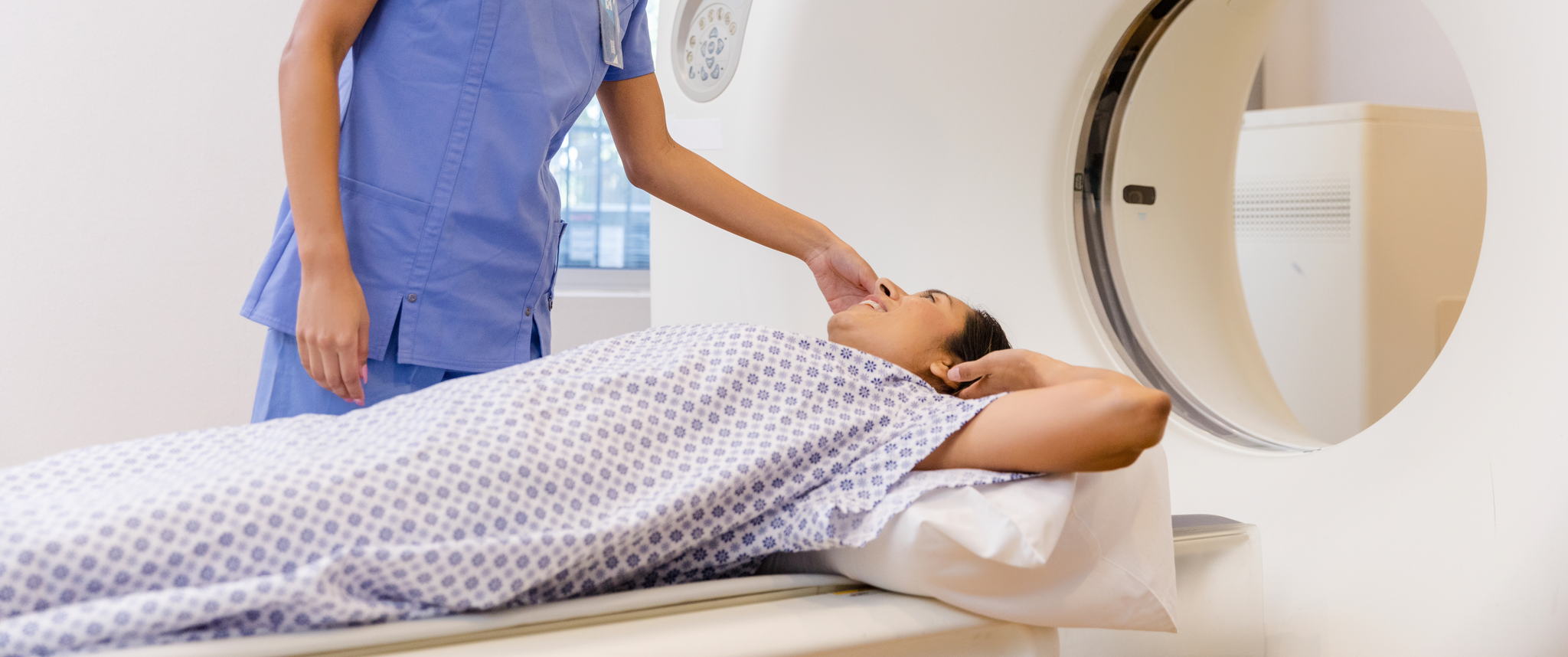 A female health care provider in scrubs tending to a patient in a hospital gown by a MRI machine