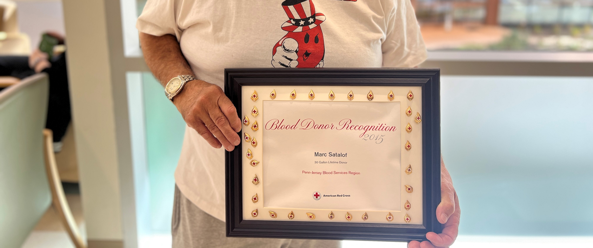 Marc Satalof, smiling, holds a framed certificate that says "Blood Donor Recognition"