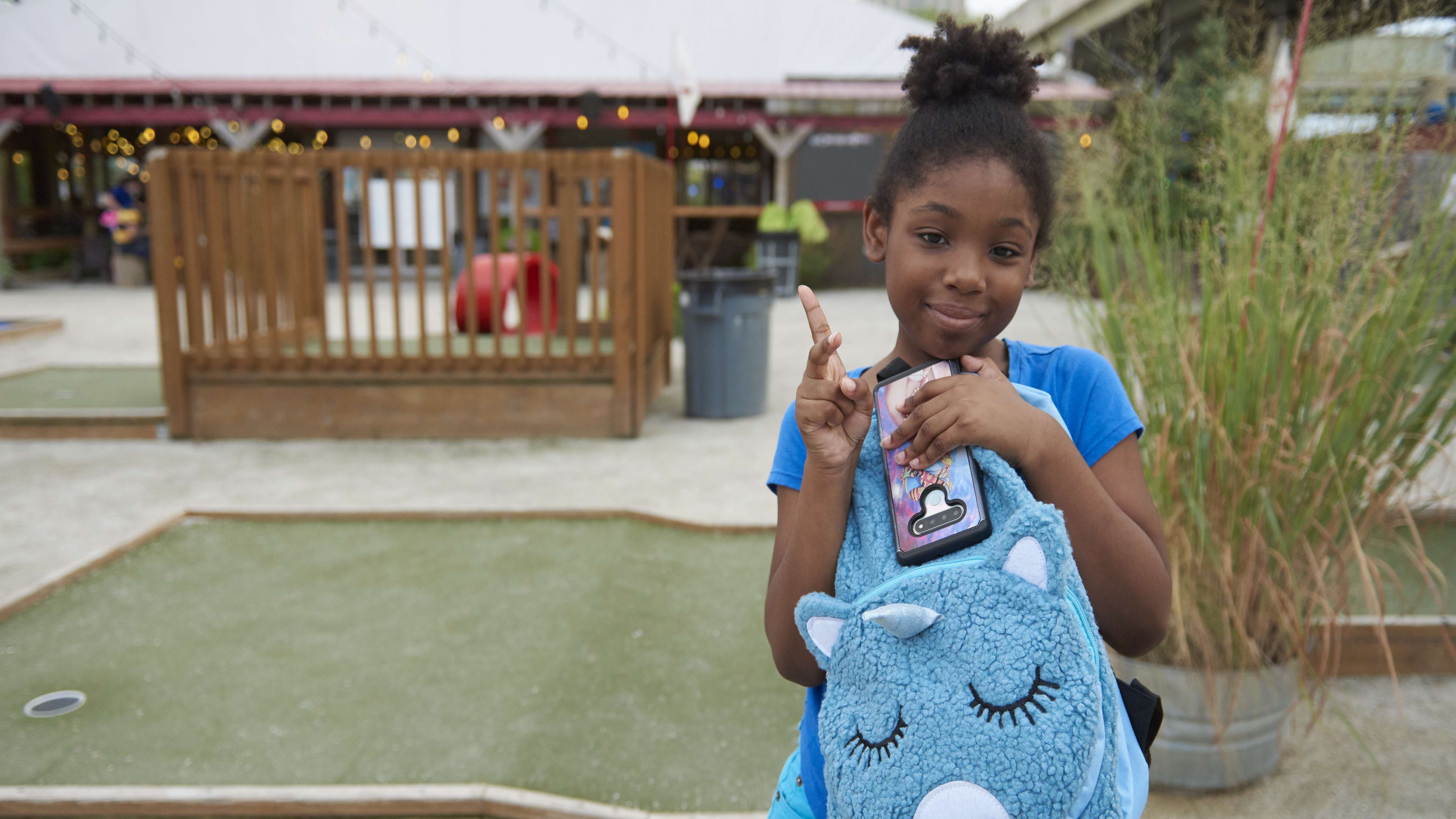 A girl standing on a mini golf course holds a blue unicorn-shaped backpack