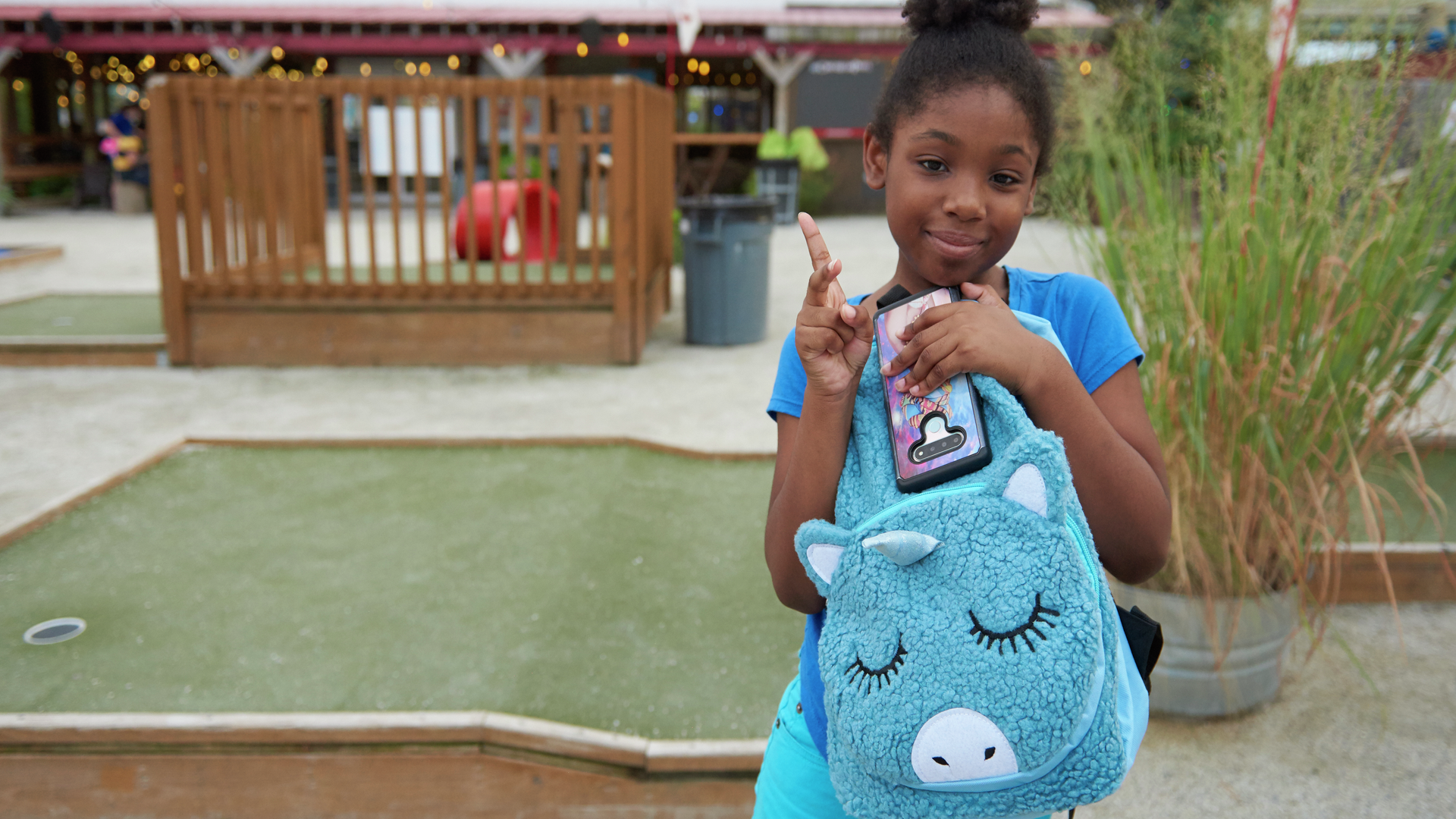 A girl standing on a mini golf course holds a blue unicorn-shaped backpack