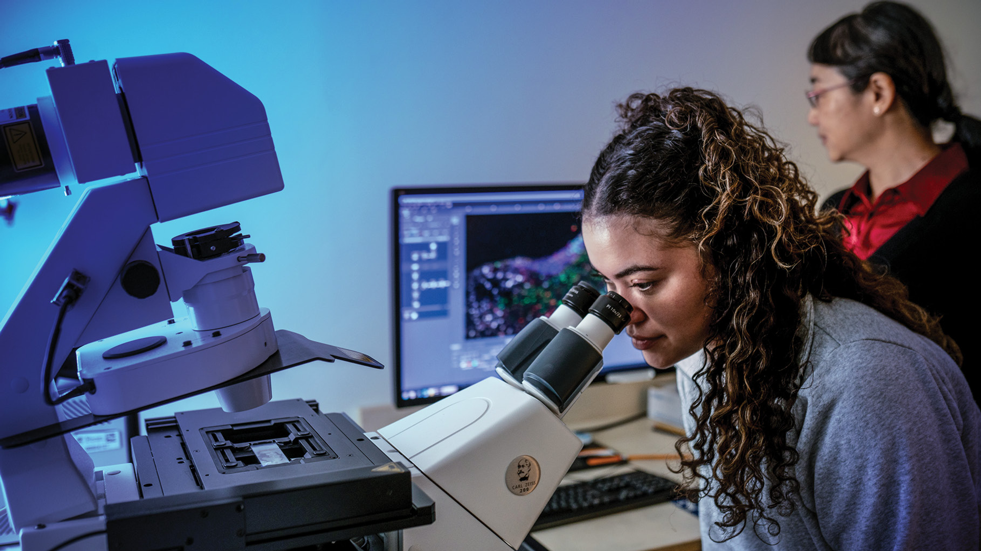 Marissa Coppin looks into a microscope while Guo-li Ming looks at the brain cell images shown on a screen beside her