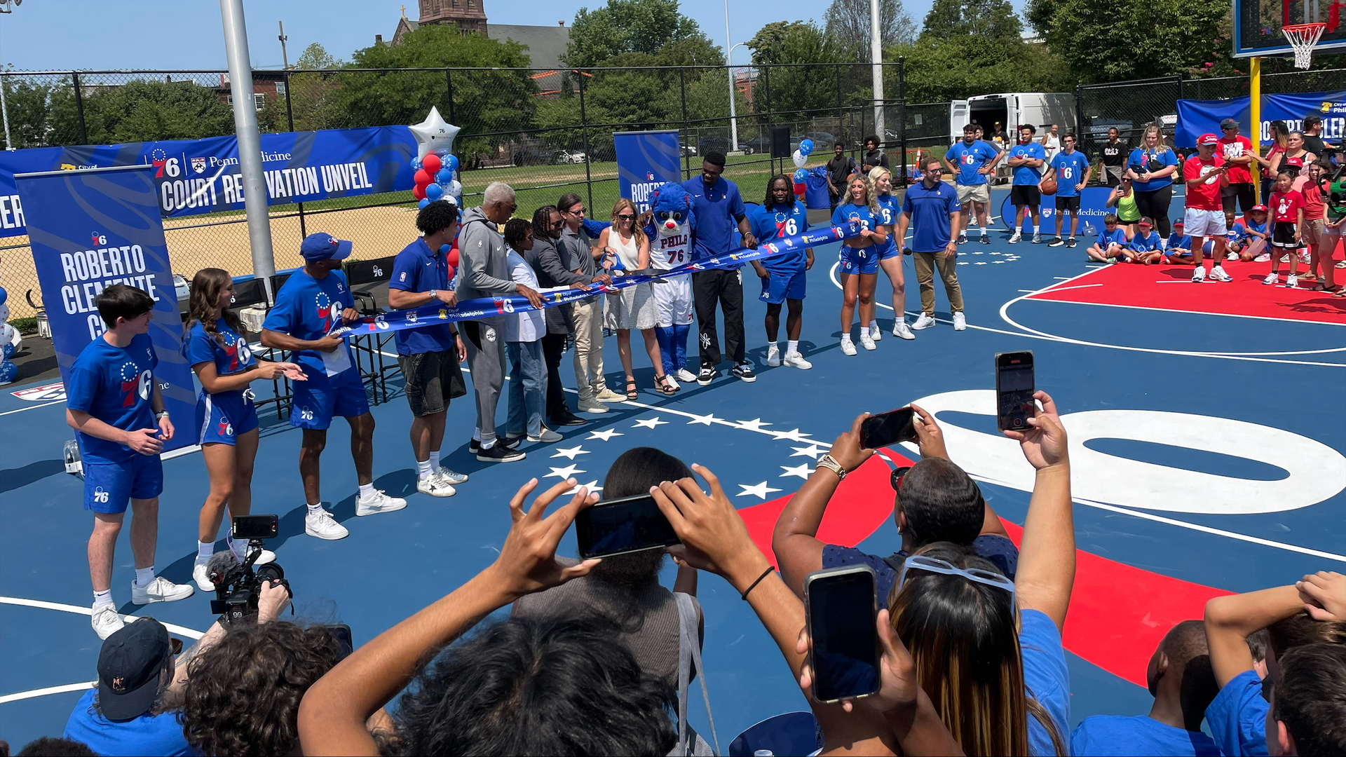 A large group of people, many dressed in blue 76ers T-shirts, and Franklin the mascot, line up poised to cut a blue ribbon while standing on a red-and-blue basketball court