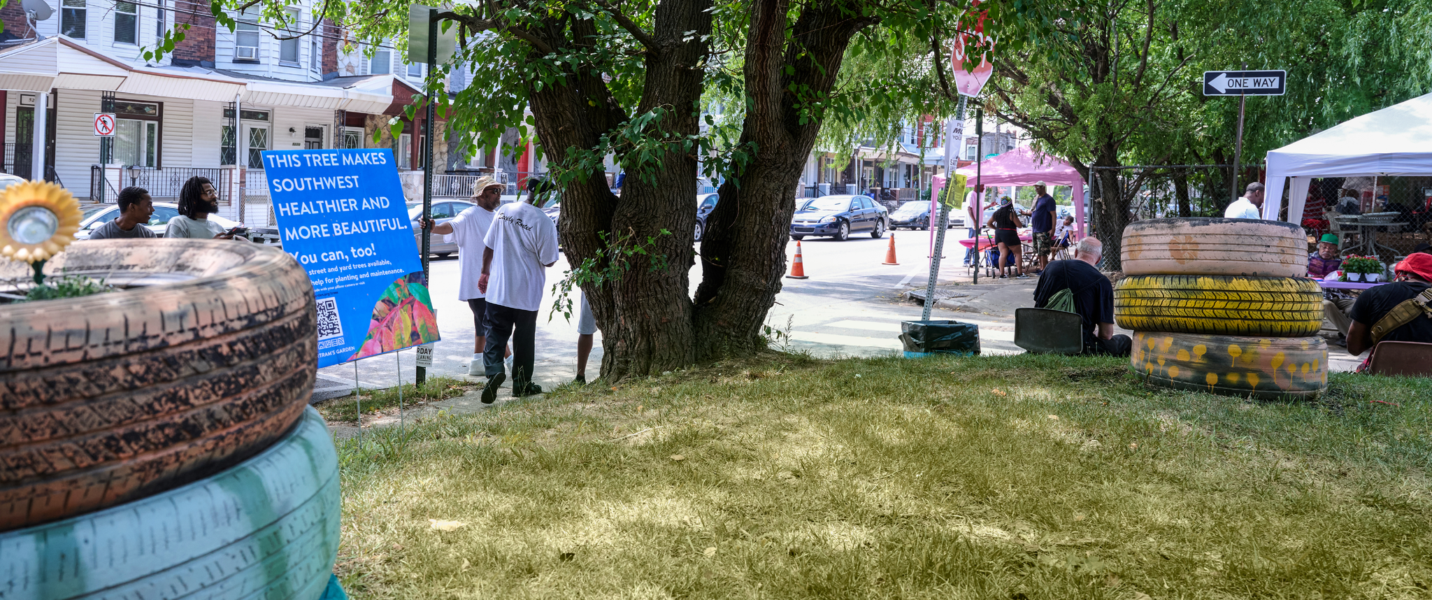 On a grassy lot on an urban street, several Black men stand beside a large tree and a sign saying This tree makes Southwest Philadelphia more beautiful