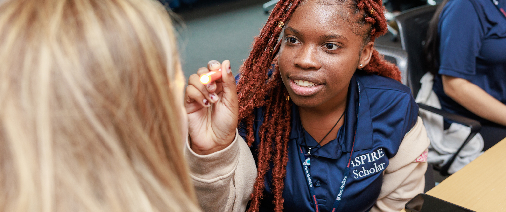 A young Black woman wearing a shirt with an ASPIRE logo holds up a light to examine someone's eyes