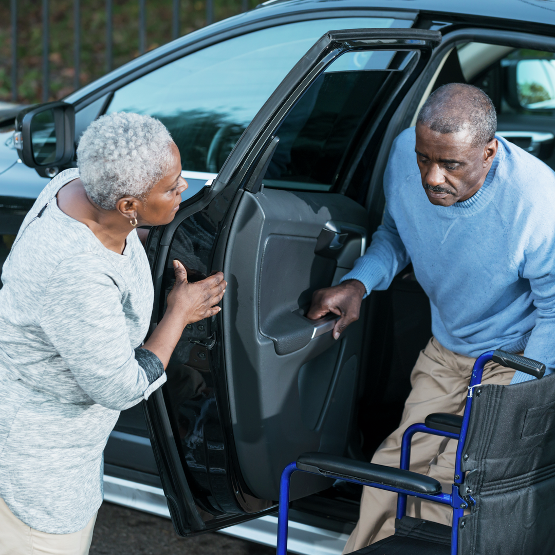 An African American senior woman helping her disabled husband climb out of a car into his wheelchair.