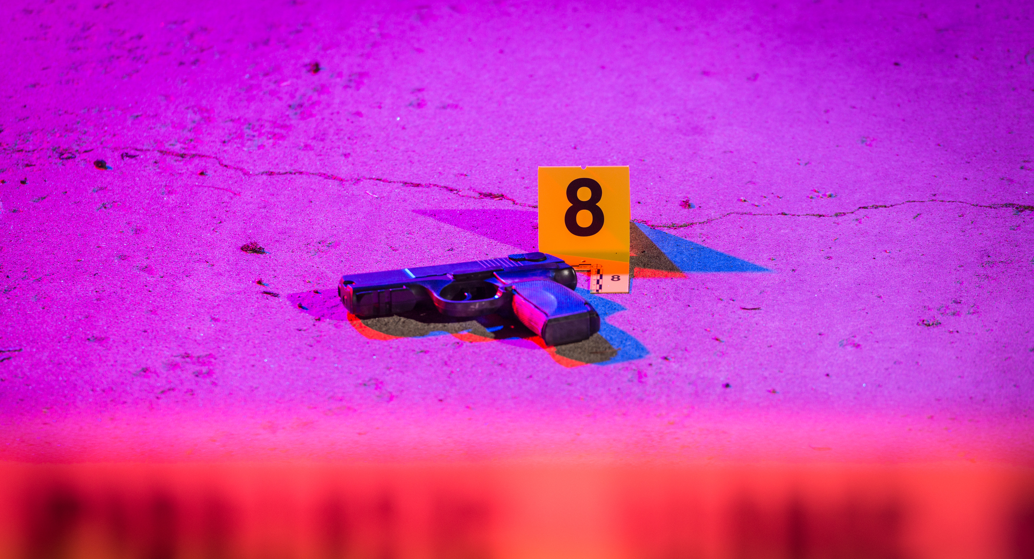 A black handgun lays on the pavement in a crime scene, marked with an evidence marker