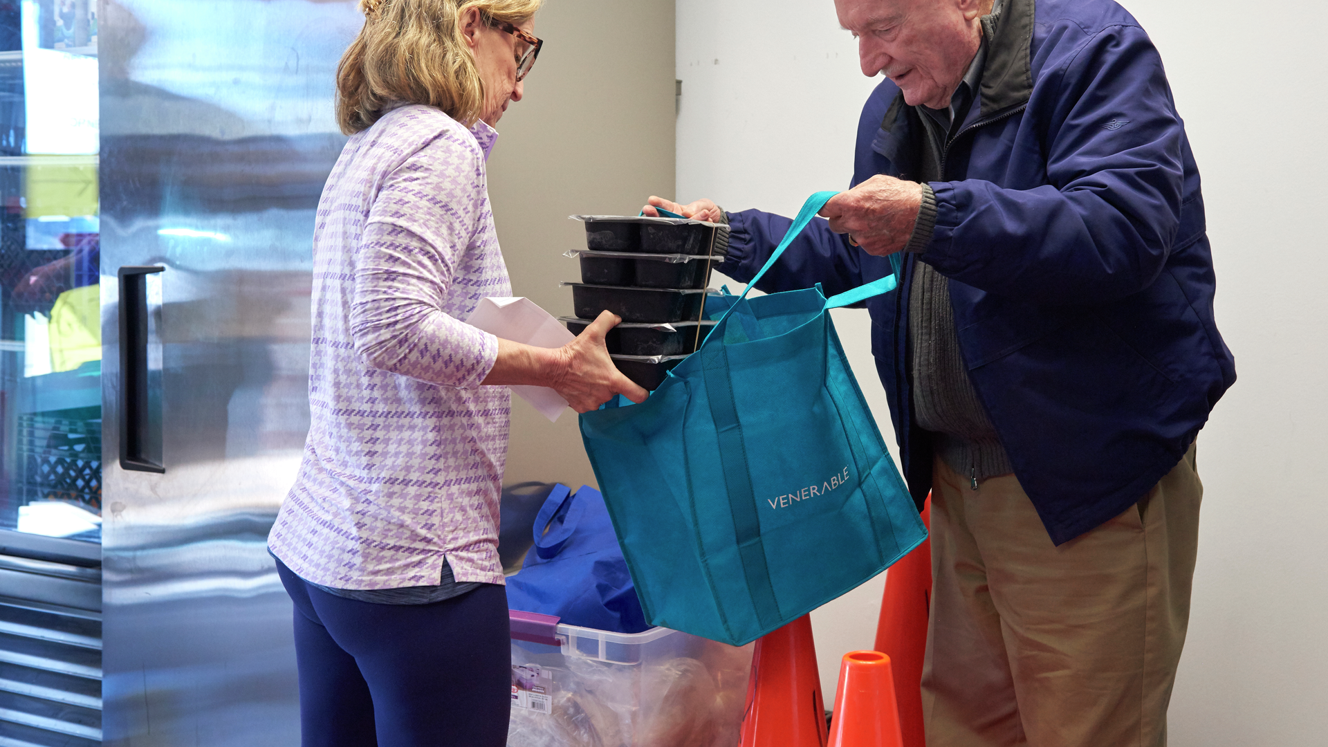 A woman places a stack of plastic food containers into a reusable shopping bag held by an older man