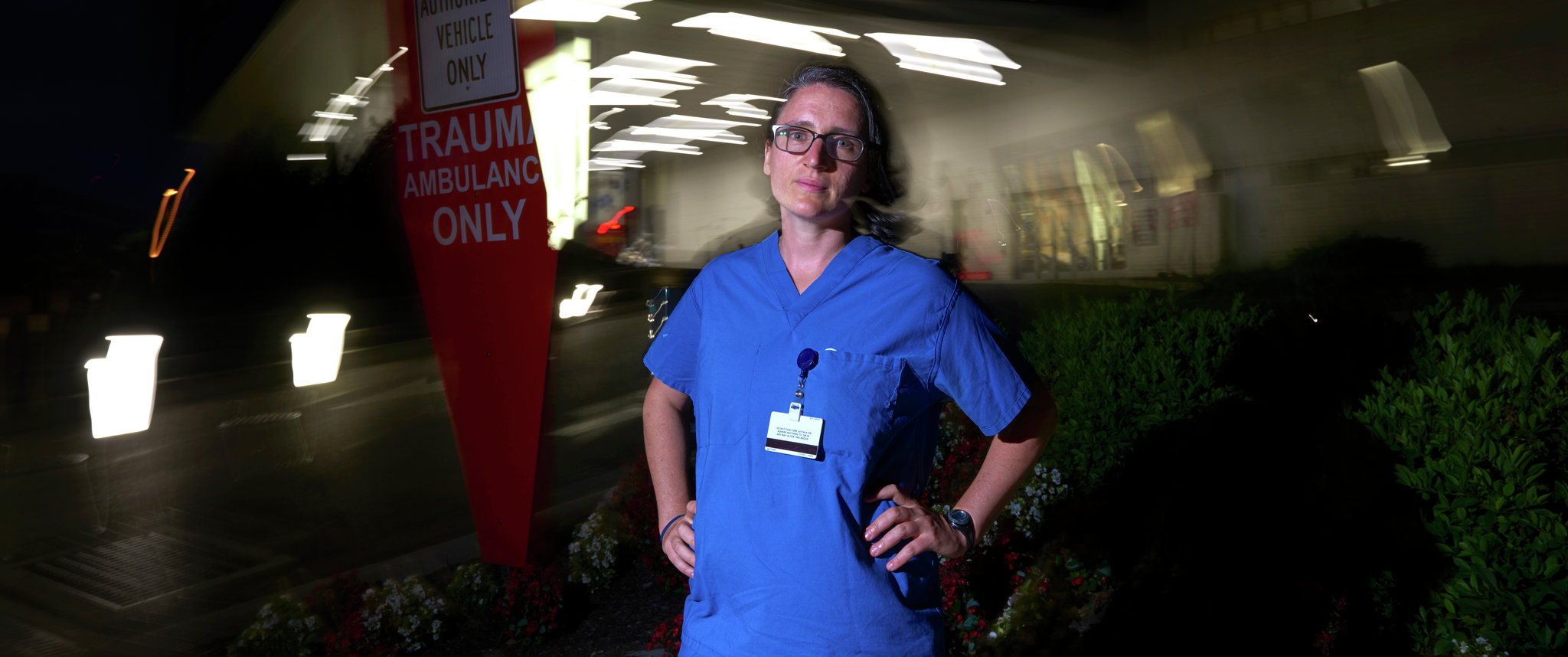 Elinore Kaufman, MD, dressed in scrubs, stands outside the Emergency/Trauma entrance of the hospital at night