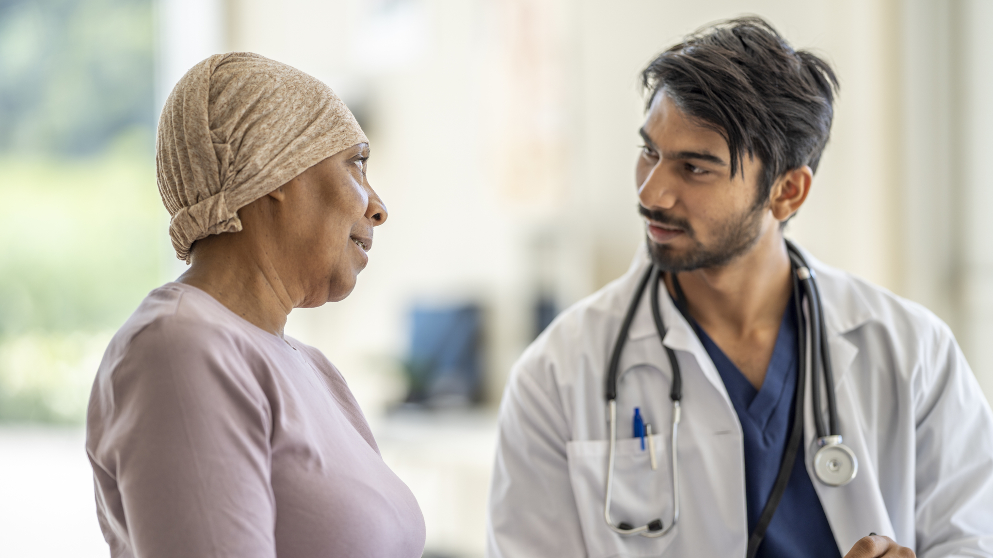 A male Oncologist of Middle Eastern decent sits with a senior patient as they discuss her plan of care. The patient is dressed casually and wearing a head scarf to keep her warm. The doctor is wearing a white lab coat and is holding a tablet as the two meet.