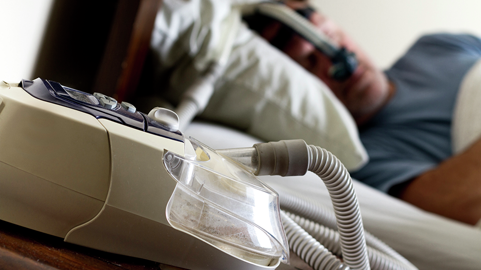 A man sleeps wearing a CPAP mask in the background; in the foreground a CPAP machine rests on a table