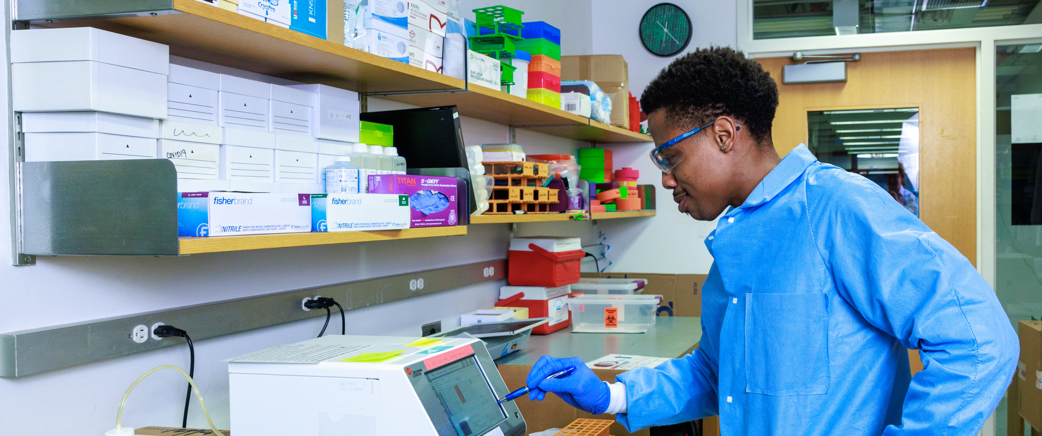 A man in a blue lab coat and safety goggles points a pen at a piece of lab equipment on a bench