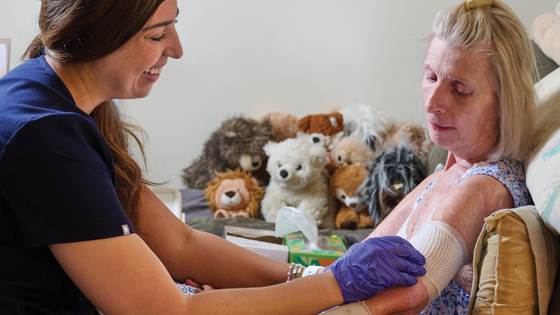 A home care nurse smiles as she checks the bandage of a female patient sitting on a couch