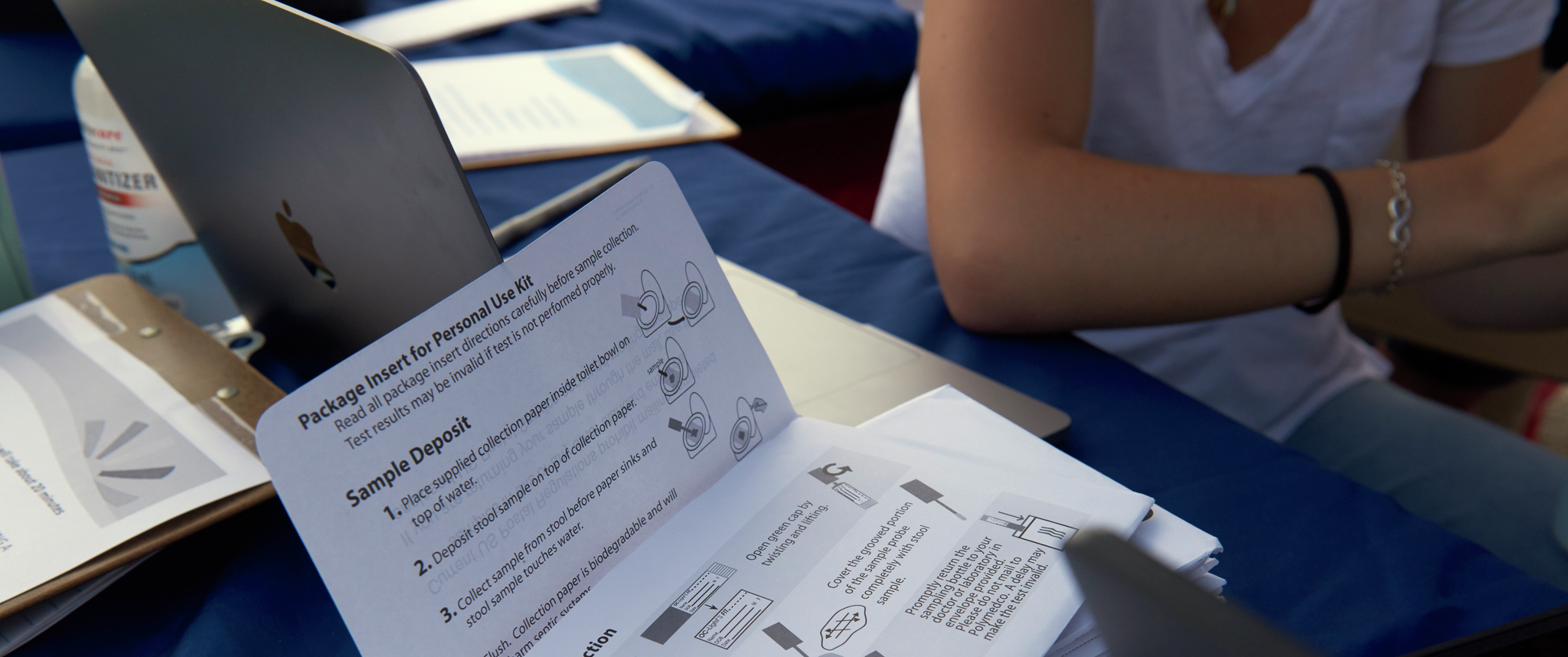 An unfolded paper on a table next to laptops contains instructions on how to use a home fecal test kit for colorectal cancer