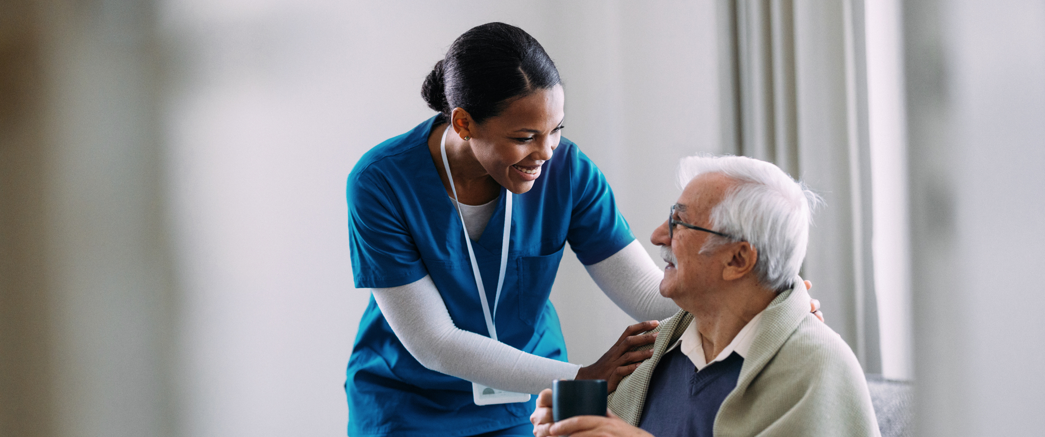 A compassionate nurse in a blue uniform helps an elderly man with a drink, creating a warm and caring atmosphere in a home setting. The scene highlights caregiving and support.