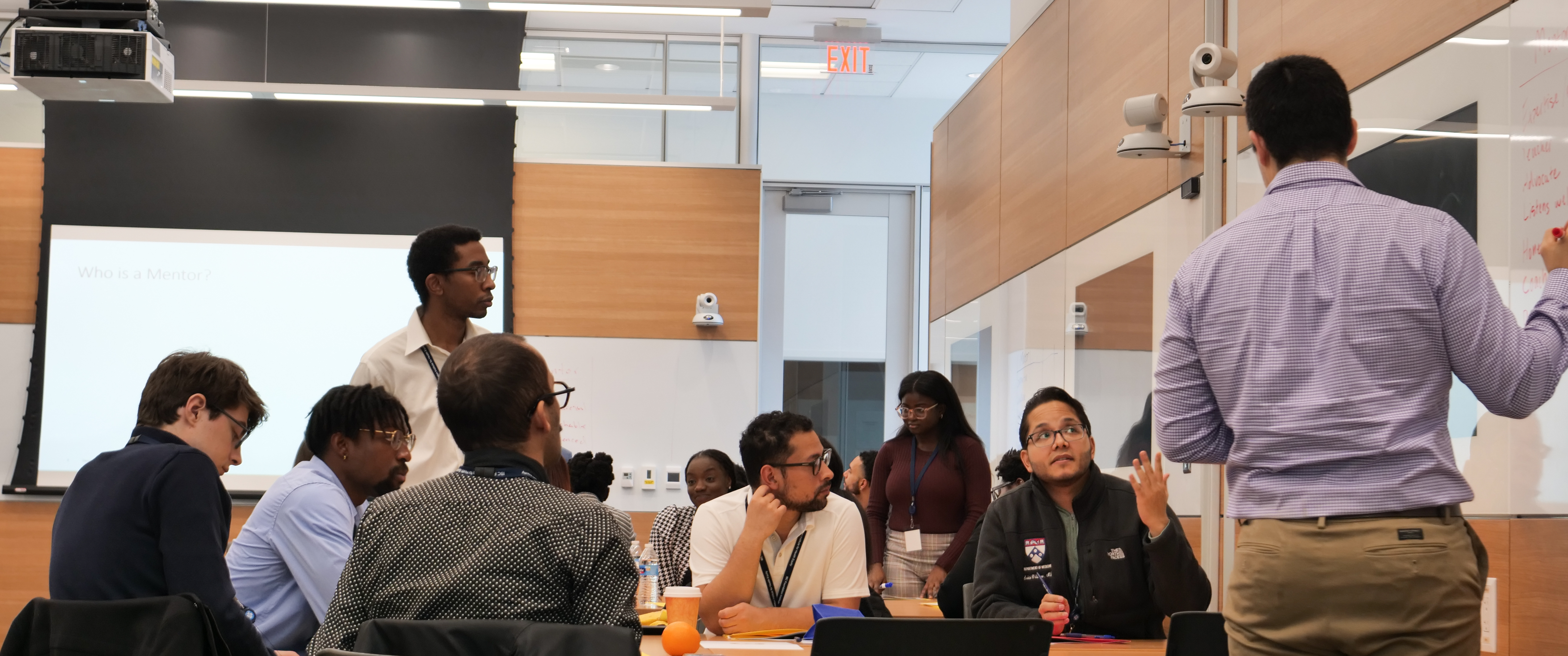 A group of Black and other underrepresented minority medical students sit in a modern classroom while one student writes on a whiteboard.