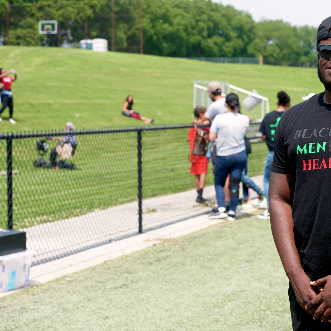 A Black man stands in a confident pose on an athletic field; his shirt says Black Mental Health