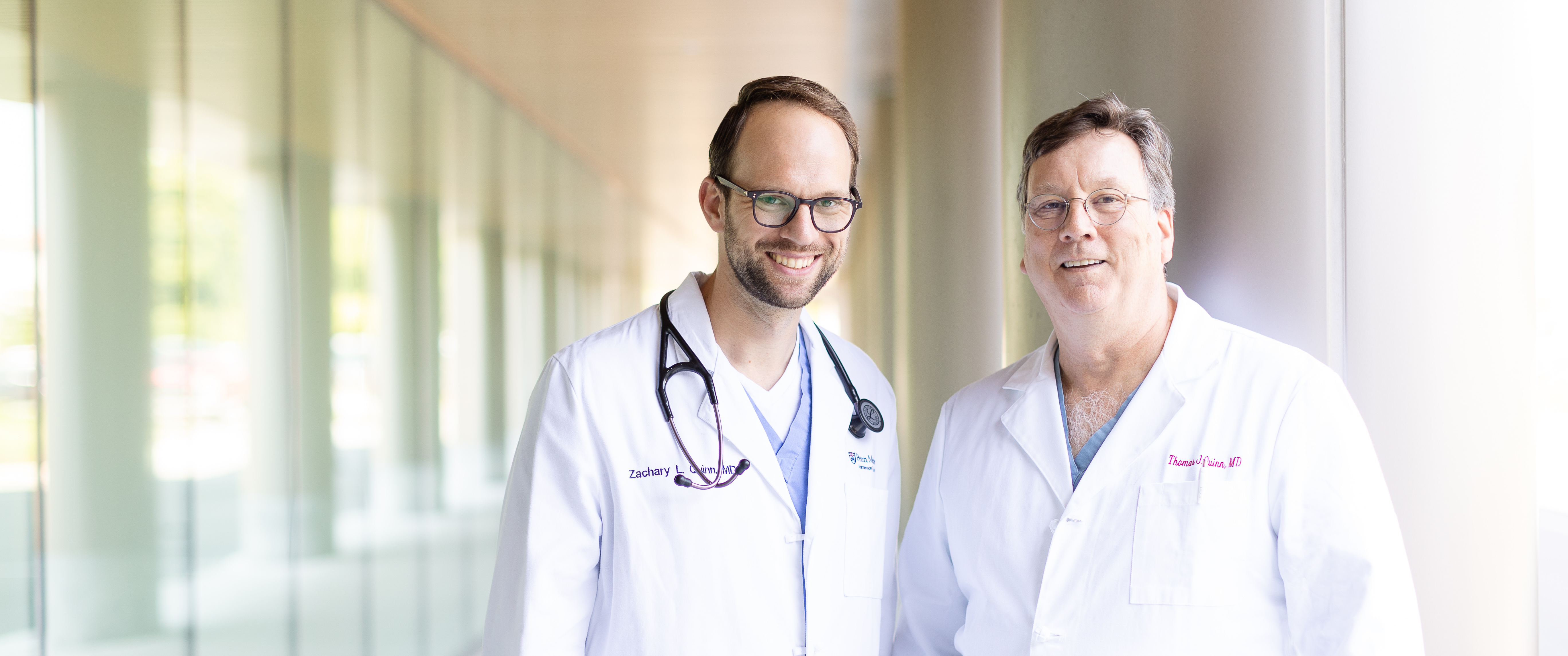 Father and son, Dr. Tom Quinn and Dr. Zack Quinn, standing together wearing physician white coats