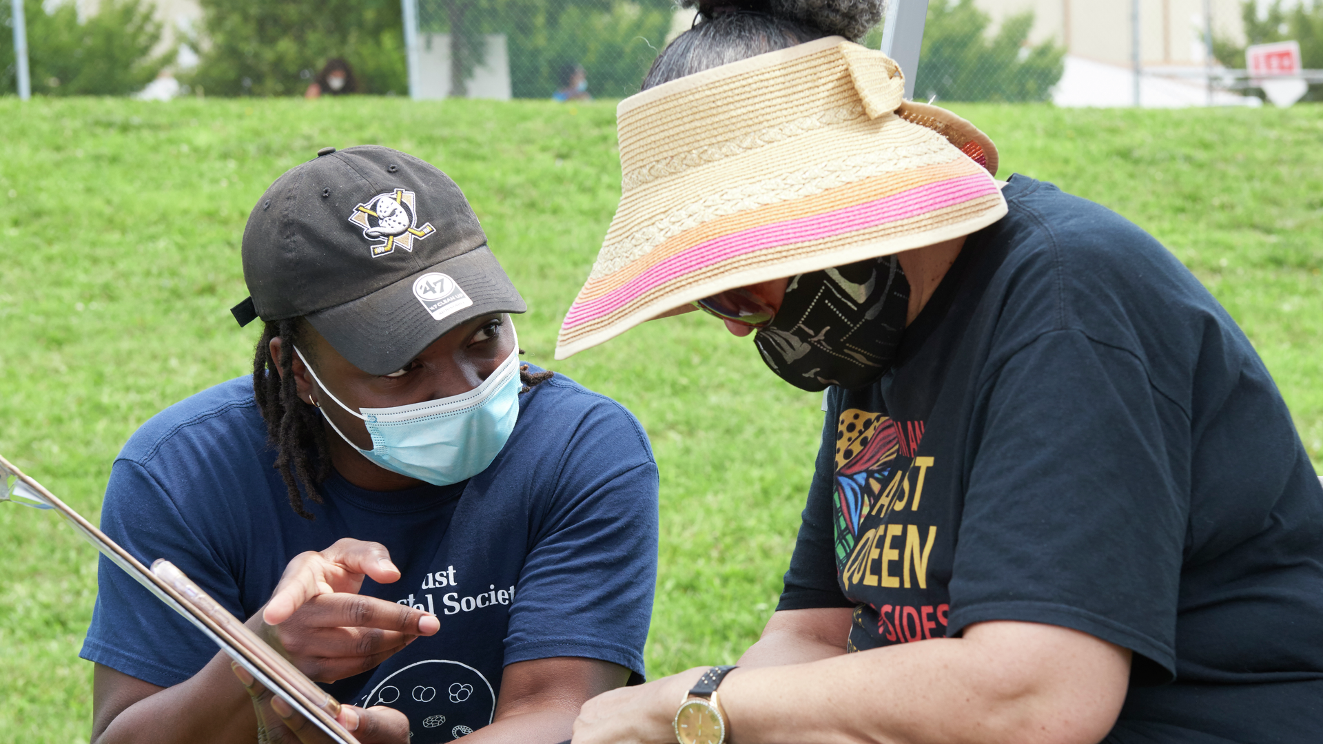 A young black man kneels beside an older woman wearing a mask and sun visor, as he holds a clipboard