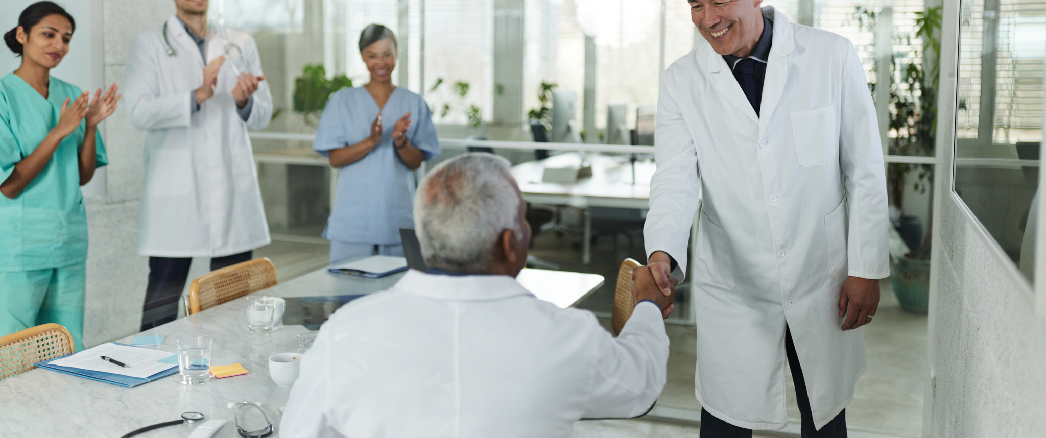 Two doctors shaking hands smiling while other medical professionals applaud 