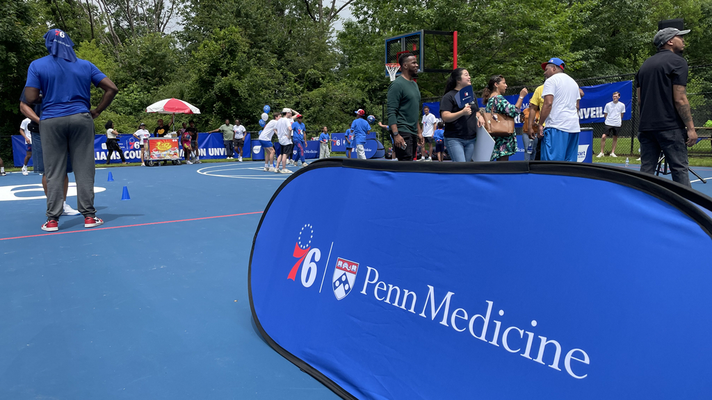 A sign with both the Philadelphia 76ers and Penn Medicine logos placed on a basketball court with children doing basketball drills in the background.
