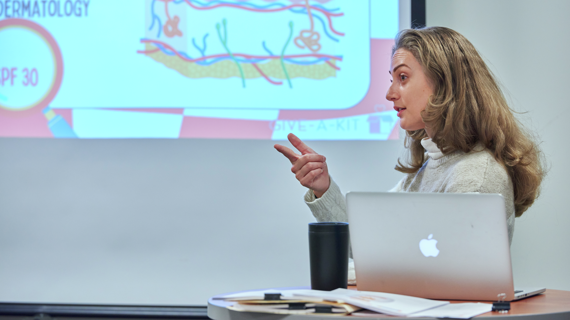 Liz Ramos stands behind a laptop, with a projected image on the wall behind her