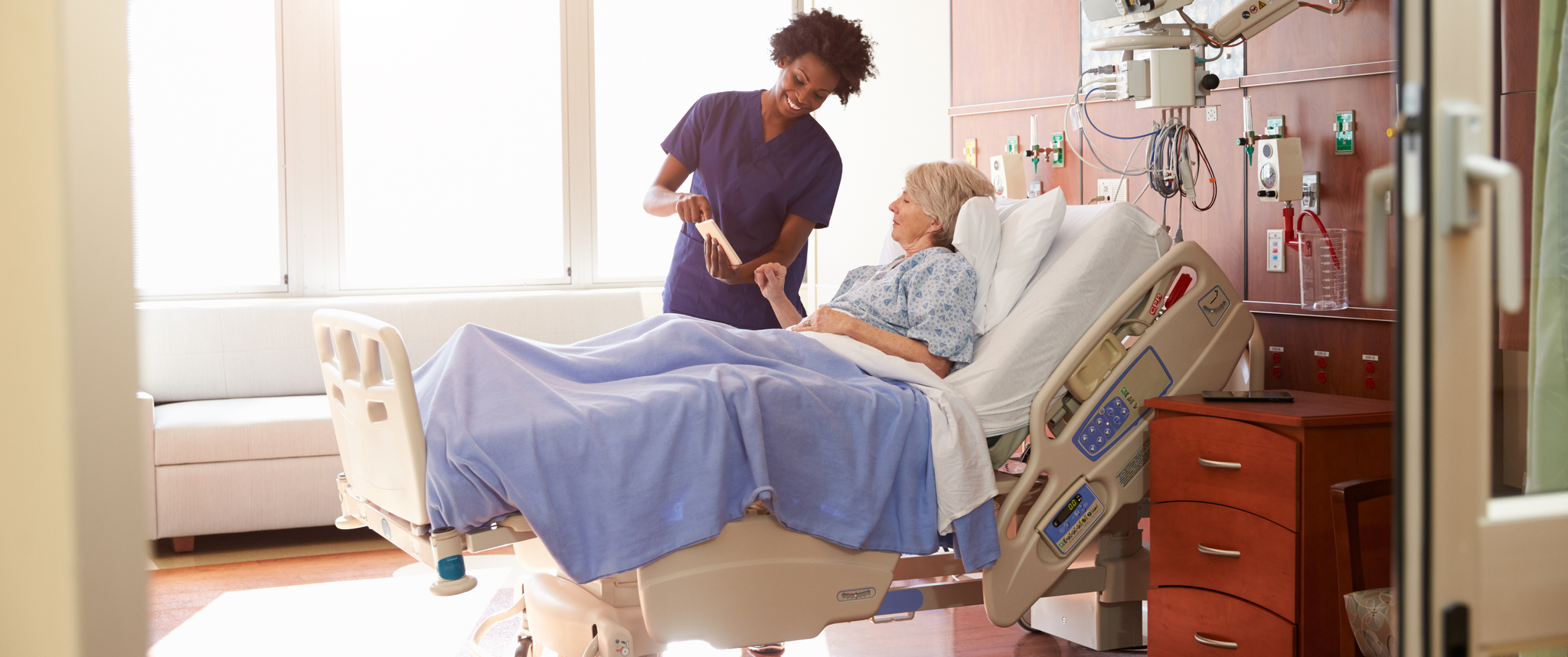 Nurse speaking with patient laying in hospital bed while showing the screen of a tablet.