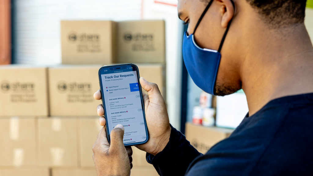 A Black man wearing a face mask looks at a smartphone screen displaying a list of food delivery requests