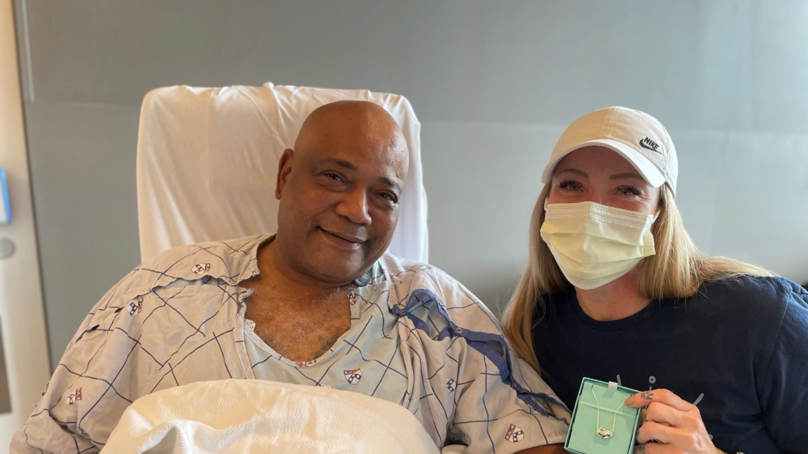  Craig Harris, a Black man in a hospital gown, smiles as a masked Morgan Slaughter leans toward him holding a heart-shaped locket