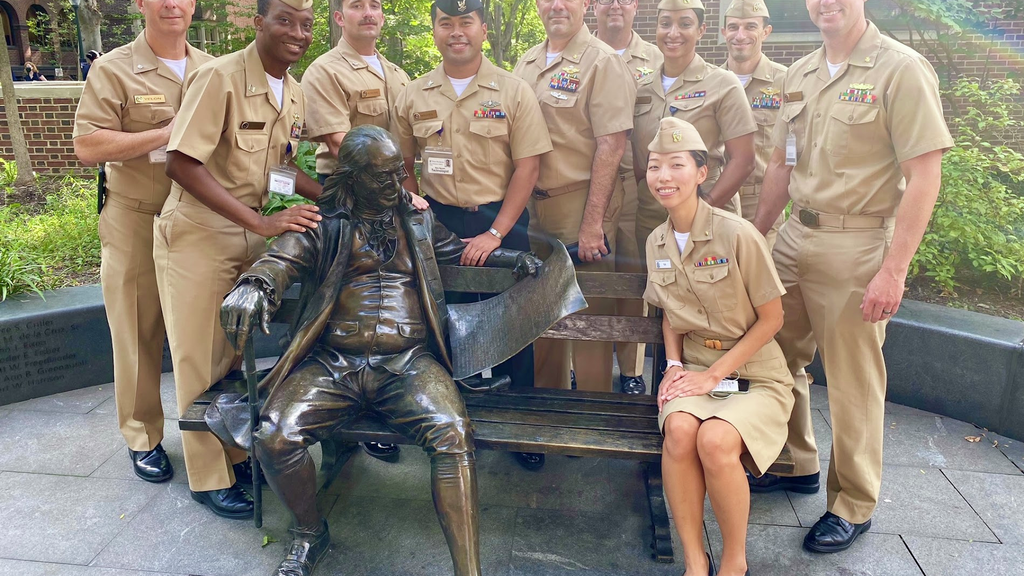 A group of Navy personnel in uniform sitting with a Ben Franklin statue on a bench