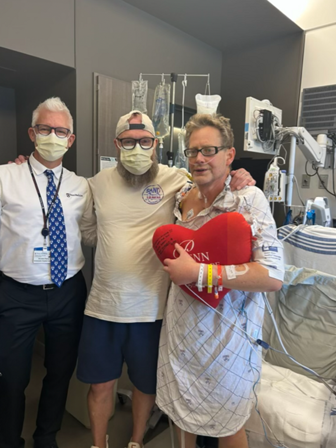 Bill Curtis, holding a heart-shaped pillow, standing with doctors and family at the Hospital of the University of Pennsylvania, post heart surgery