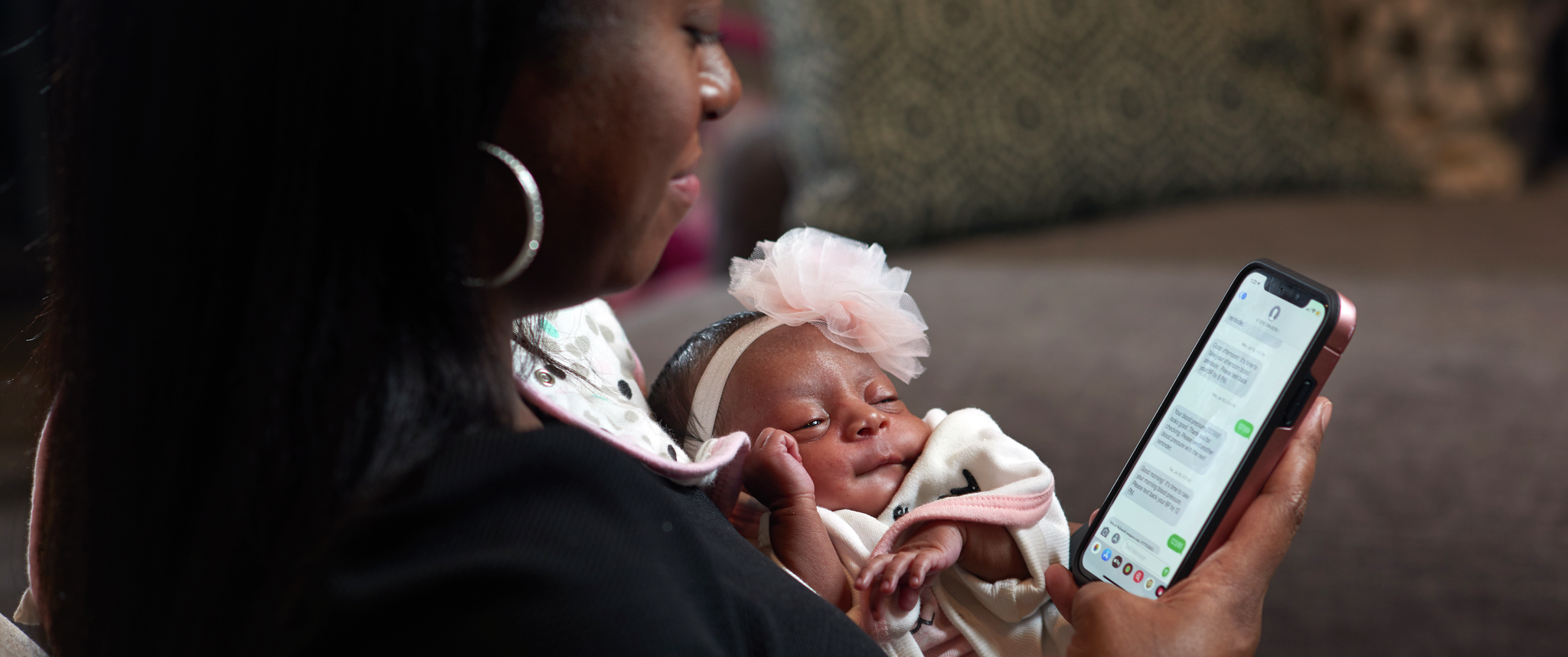 Karimah Ferguson, a young Black mother, holds her adorable baby while looking at text messages about blood pressure readings on her phone