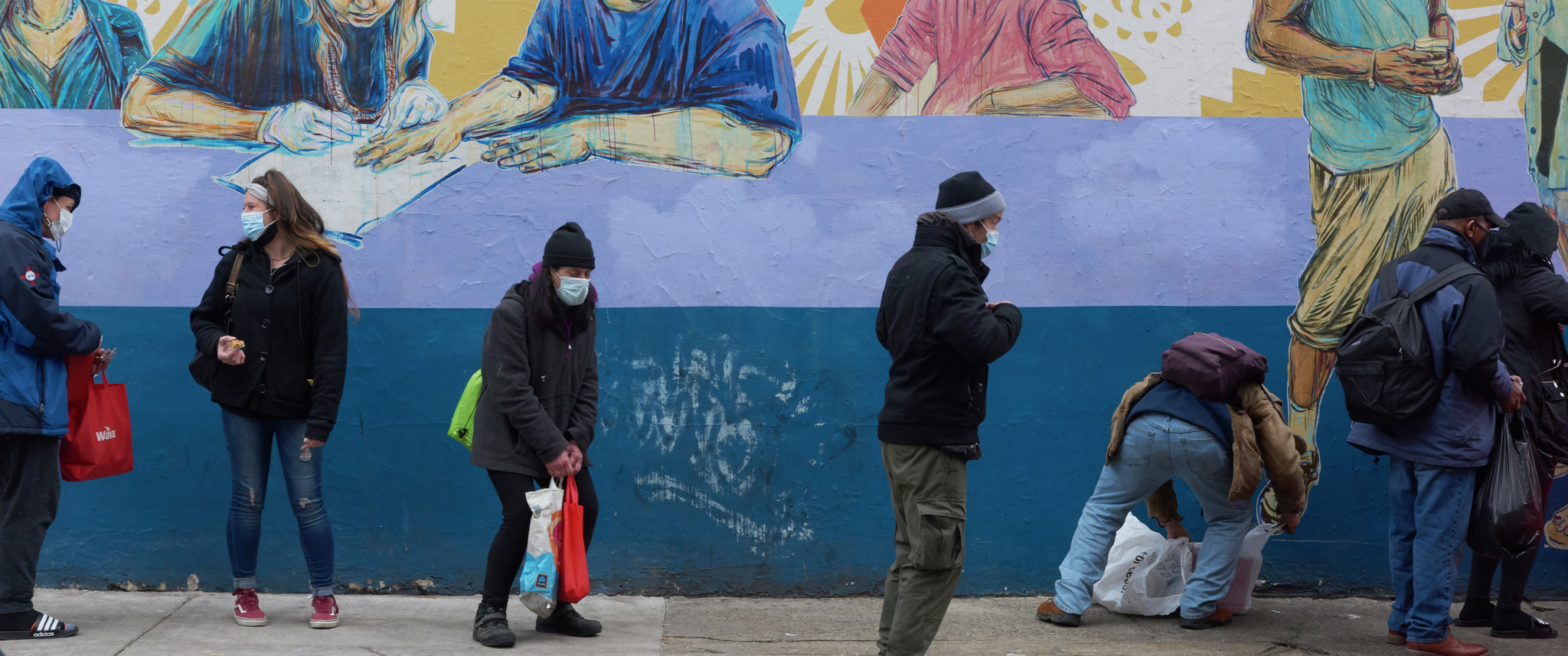 People dressed in heavy coats and face masks stand in a line in front of a wall with a colorful mural with bits of graffiti
