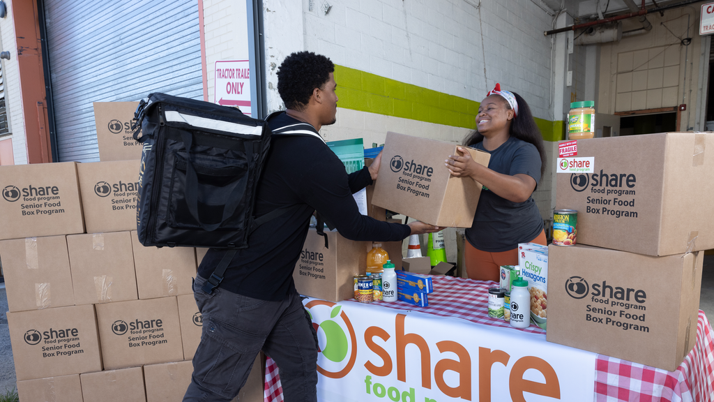 A woman standing at a table covered with food products and boxes hands a large carton to a man with a food delivery backpack
