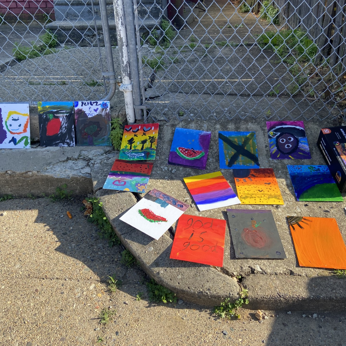 An array of small, painted canvases drying on a sidewalk next to a box of acrylic paint