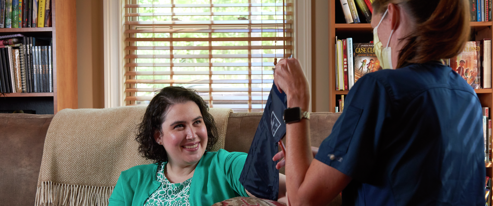 Lauren Magaziner sits on a couch at home while Erin McCarthy, RN, NP, removes a blood pressure cuff