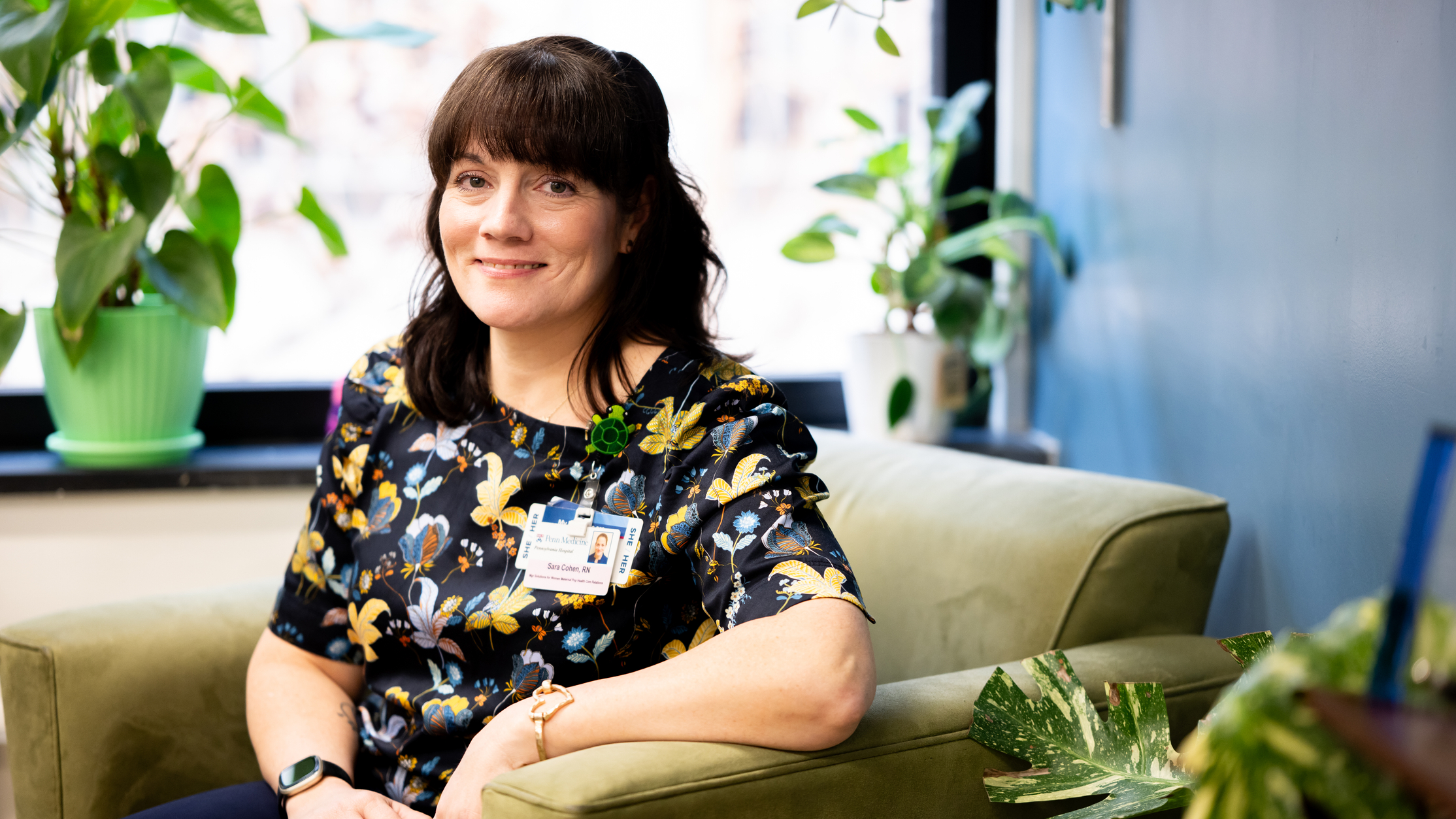 Sara Cohen sitting in a chair by a window in her office, surrounded by potted plants