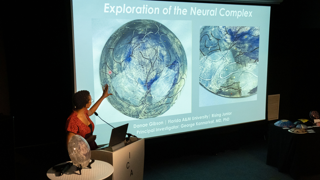 A student presenter shares their artwork during a symposium, gesturing toward the screen.