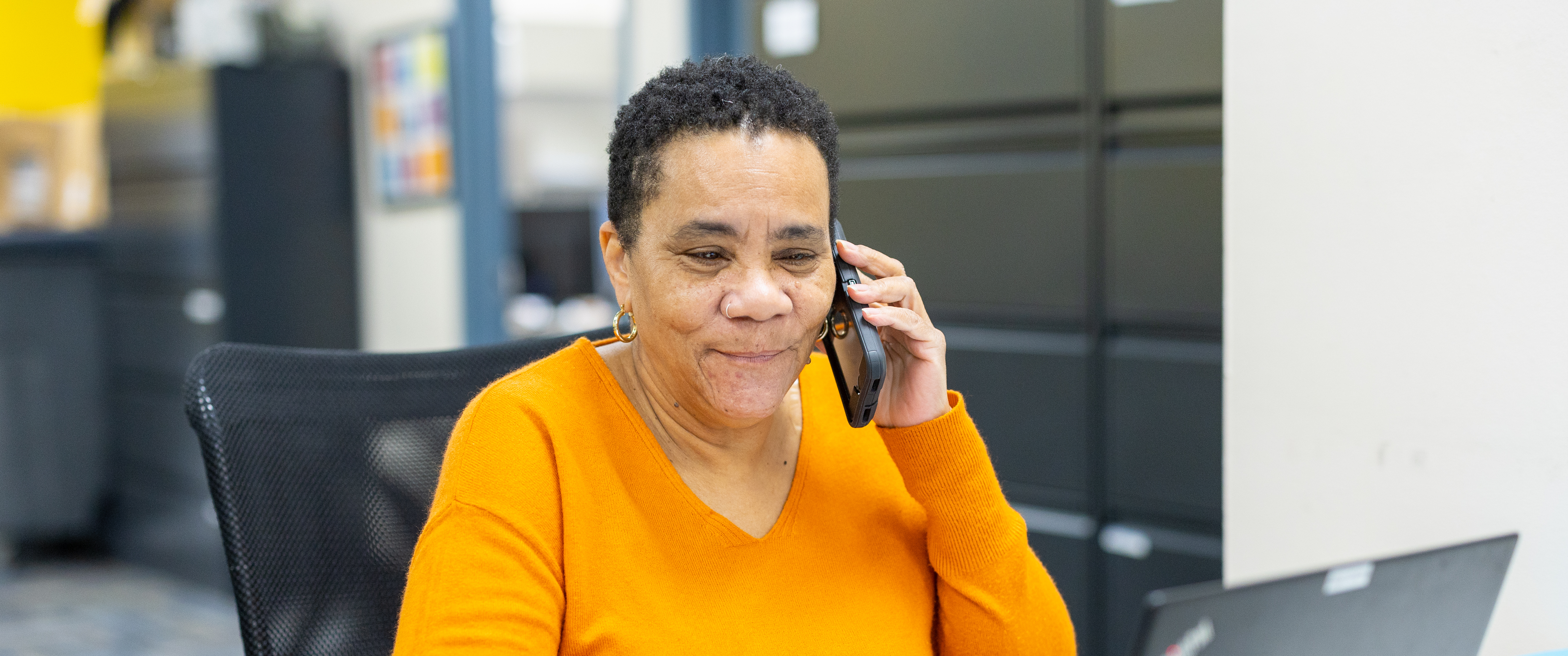Norma Gerald sits at a desk, smiling, looking at a laptop and talking on the phone