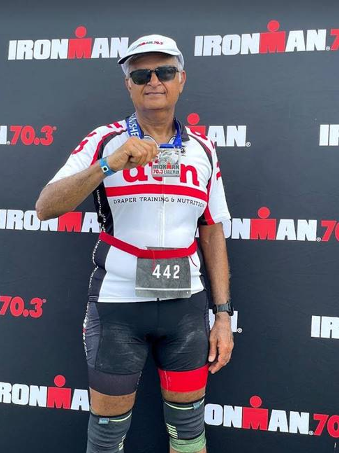 Sanjay Shah stands in front of an Iron Man 70.3 poster after a race and holds a medal for completing the triathlon