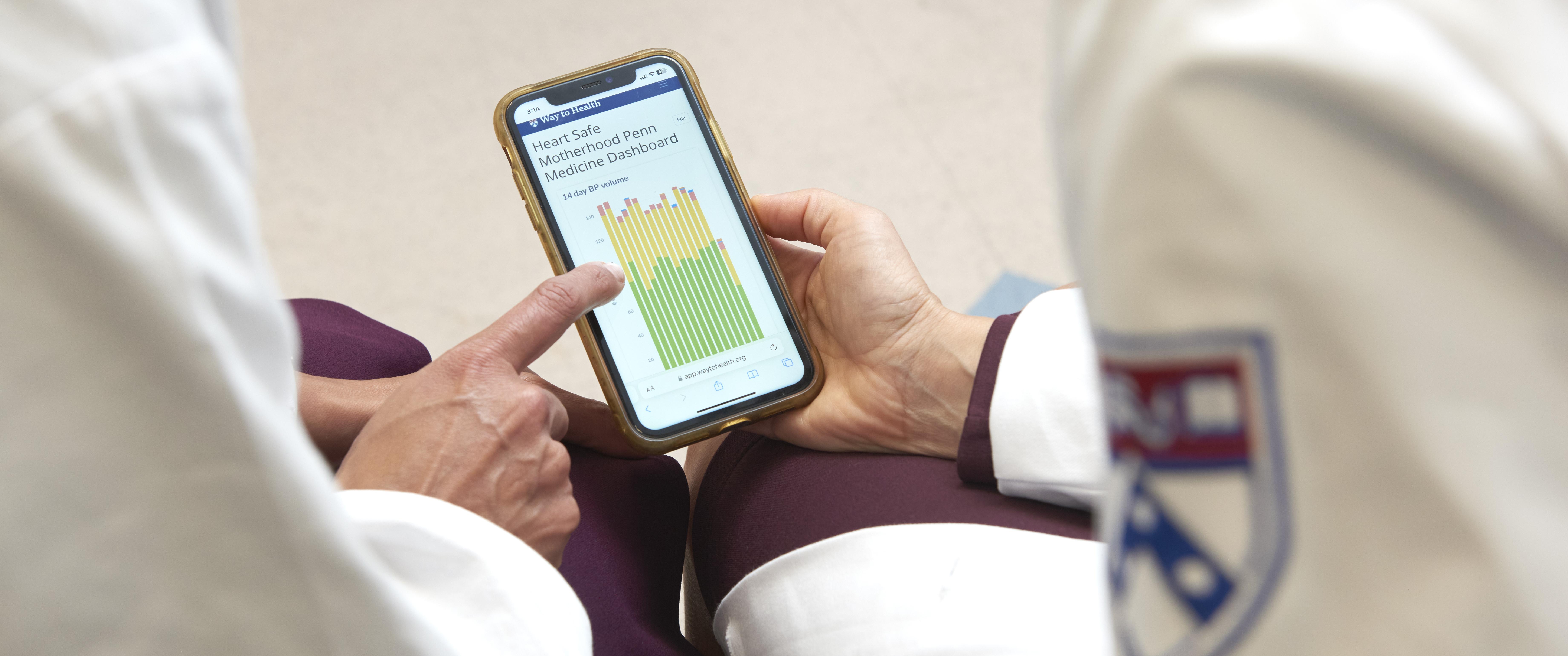 Two women's hands hold, and one points at, an iPhone showing a dashboard of data about Heart Safe Motherhood