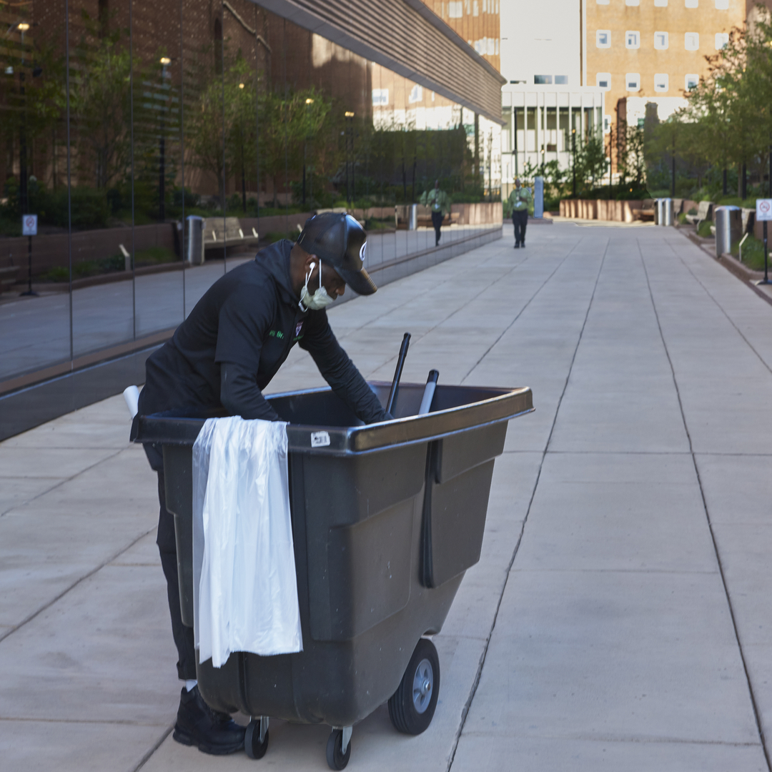 An environmental services worker with a large rolling bin stands in the middle of a wide concrete walkway with the Pavilion at his side and other HUP buildings in the background