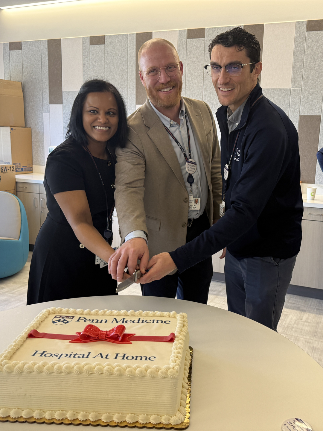 Robert Burke, Sabastian Ramagnano, and Felicia D’Souza hold a knife above a white cake with a red ribbon of frosting and icing that reads Penn Medicine
