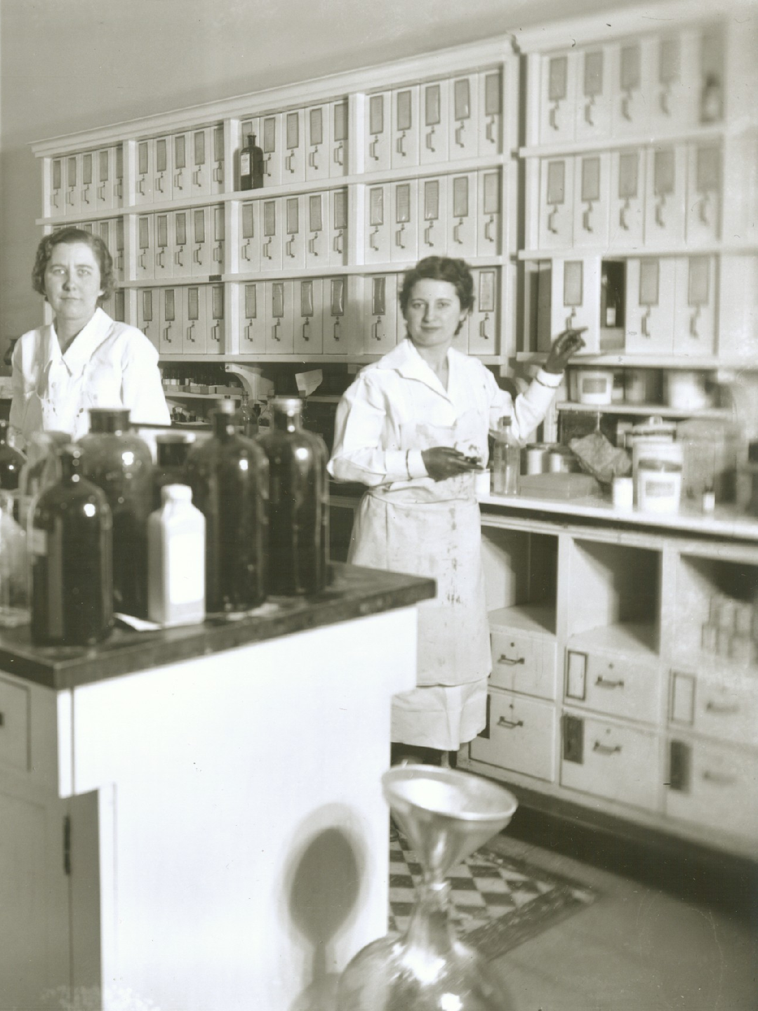 In a black and white photo, three women wearing white stand behind a large desk in the Apothecary with shelving behind them