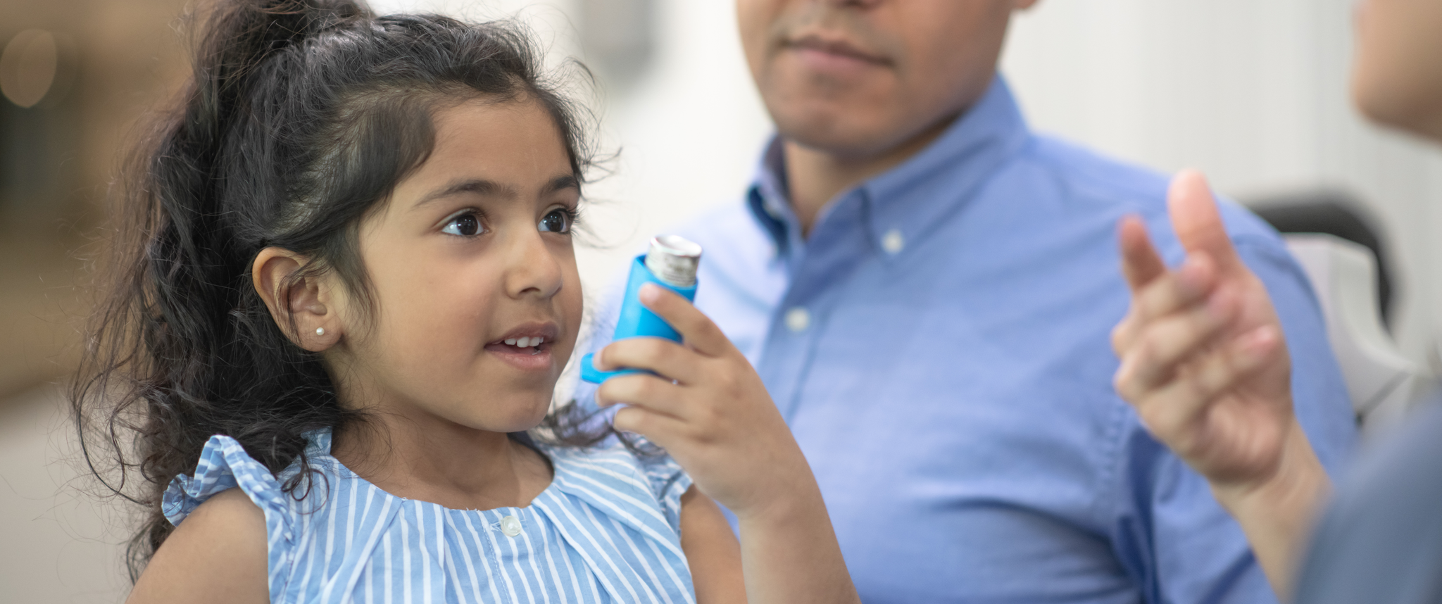 A young Hispanic girl with her father. She is holding an asthma inhaler and showing it to the provider