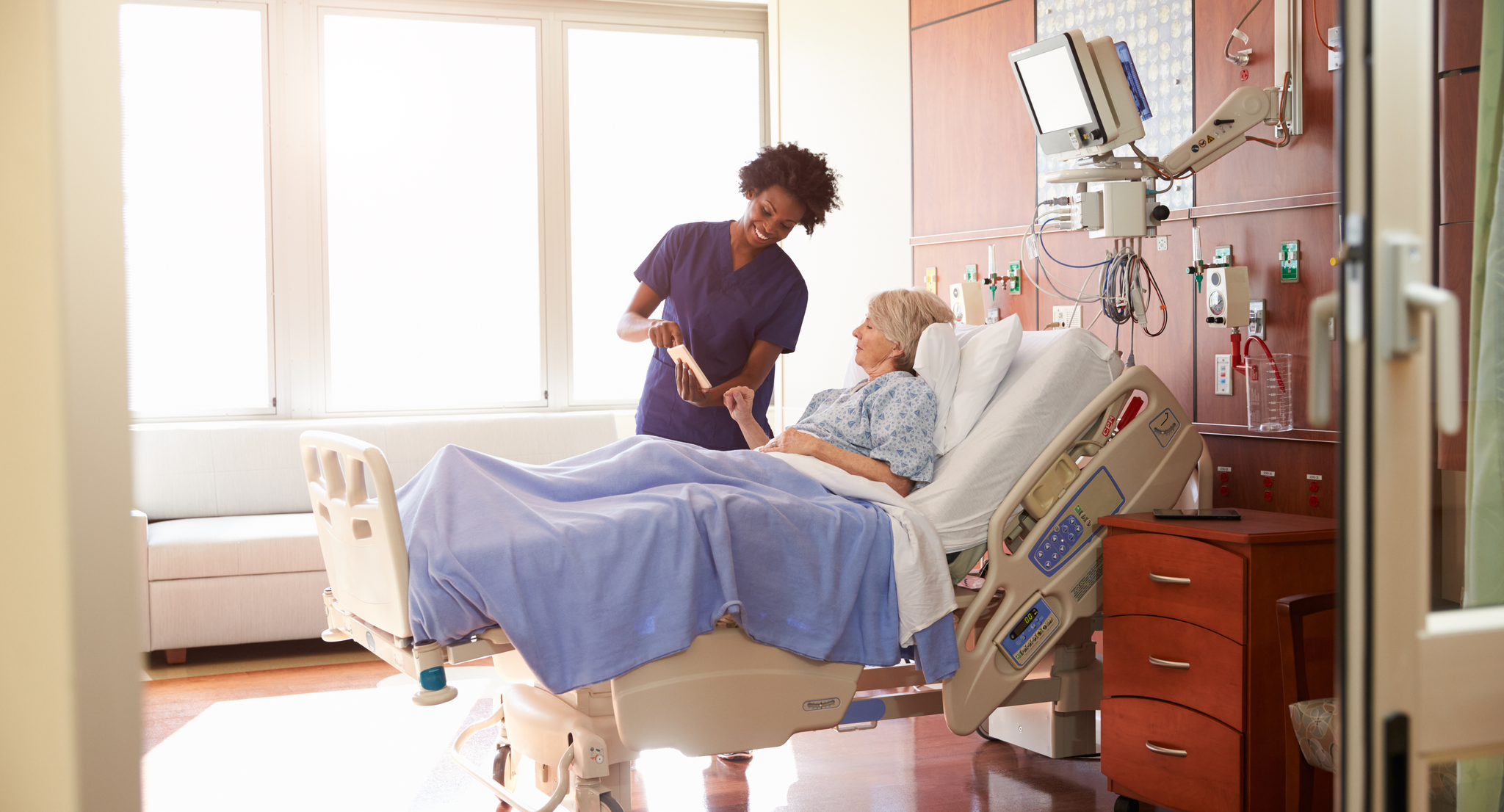 Nurse speaking with patient laying in hospital bed while showing the screen of a tablet.