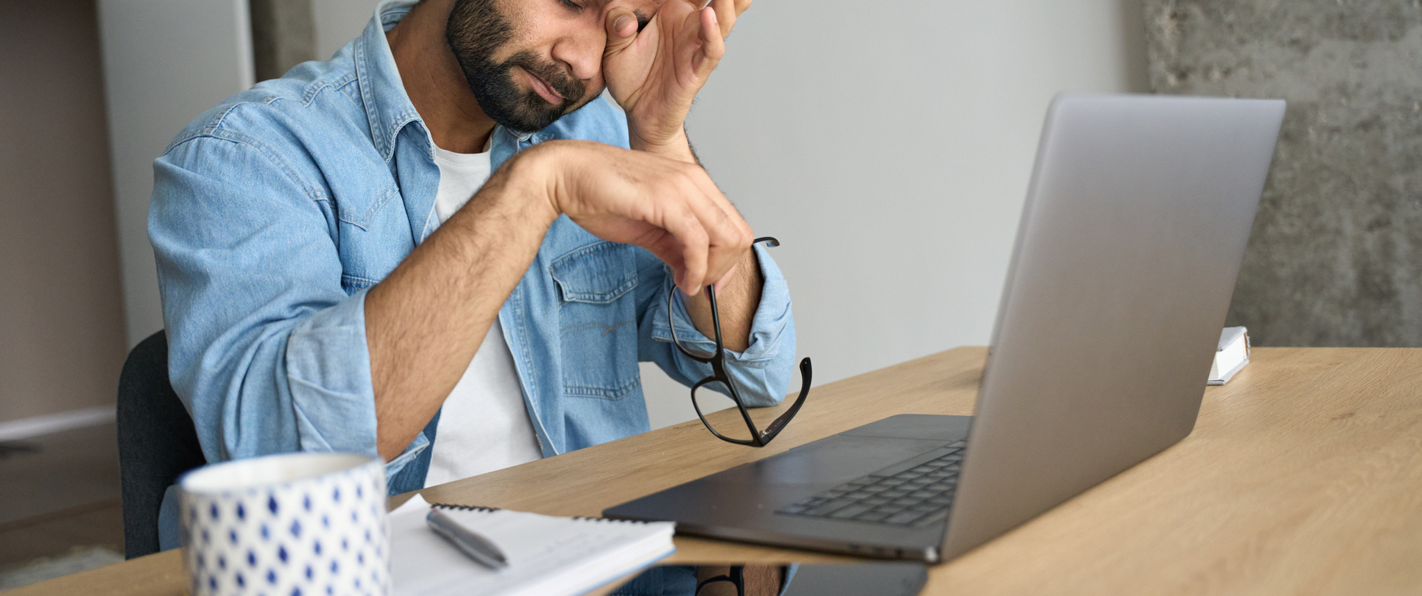 A man, with one hand covering his eyes and the other hand holding glasses, sits in front of his laptop at a desk