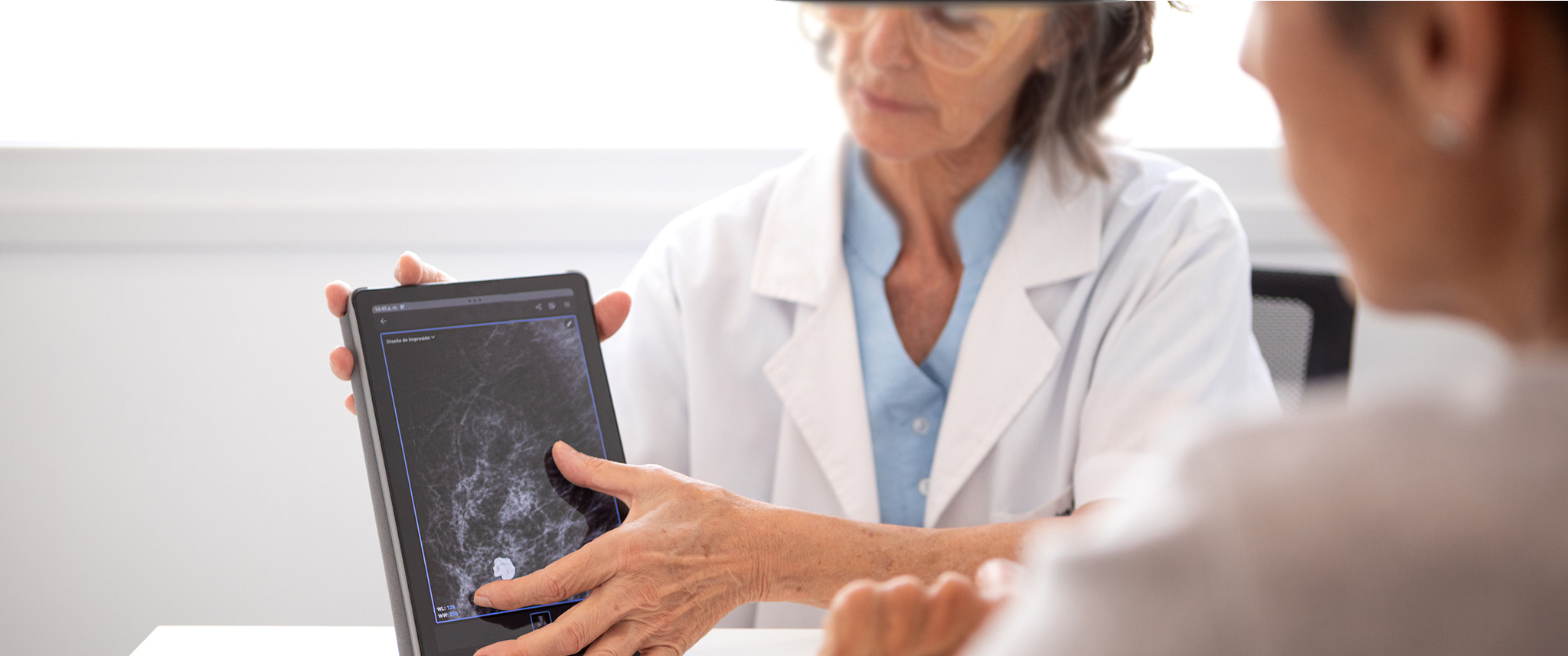 A woman doctor holds a digital device displaying a mammogram in front of an observing patient.