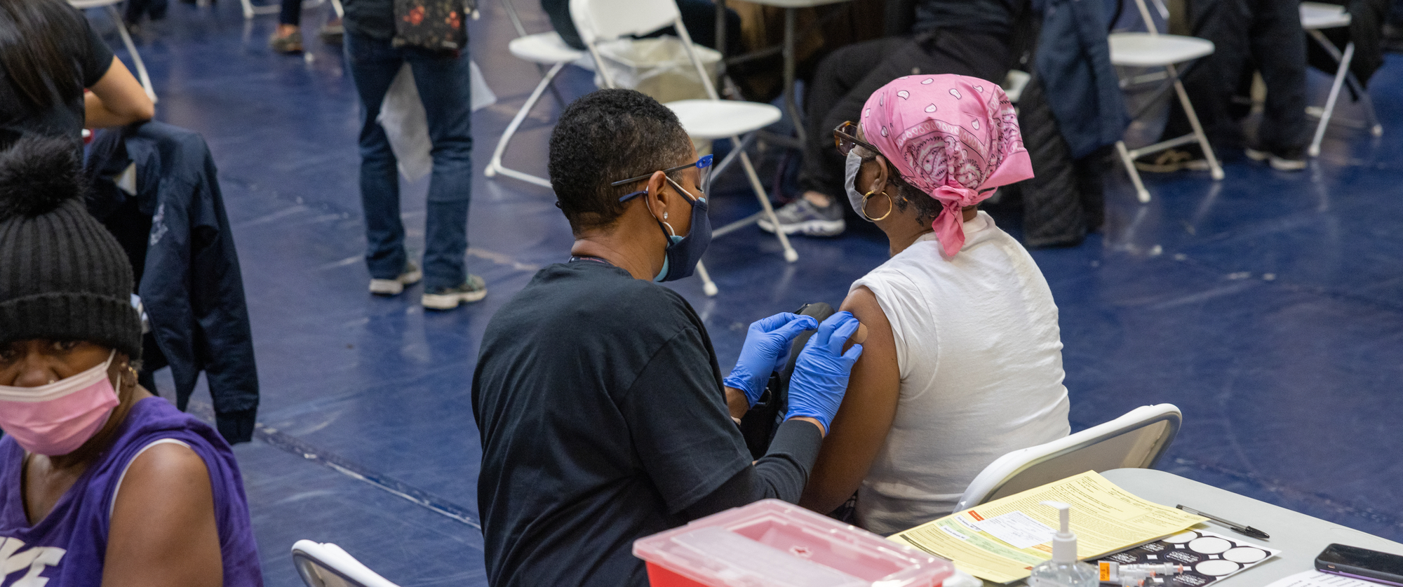 a woman wearing gloves puts a bandage on the arm of a woman who just received a COVID vaccine