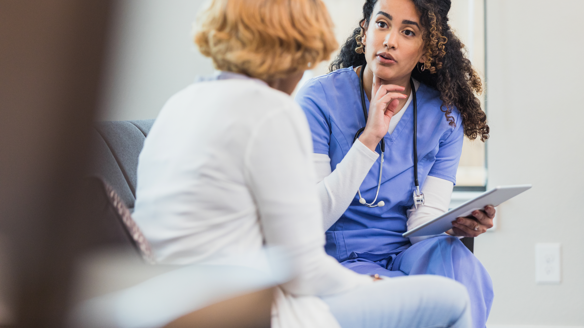 Female nurse gestures toward her neck as she asks a female patient questions about a sore throat