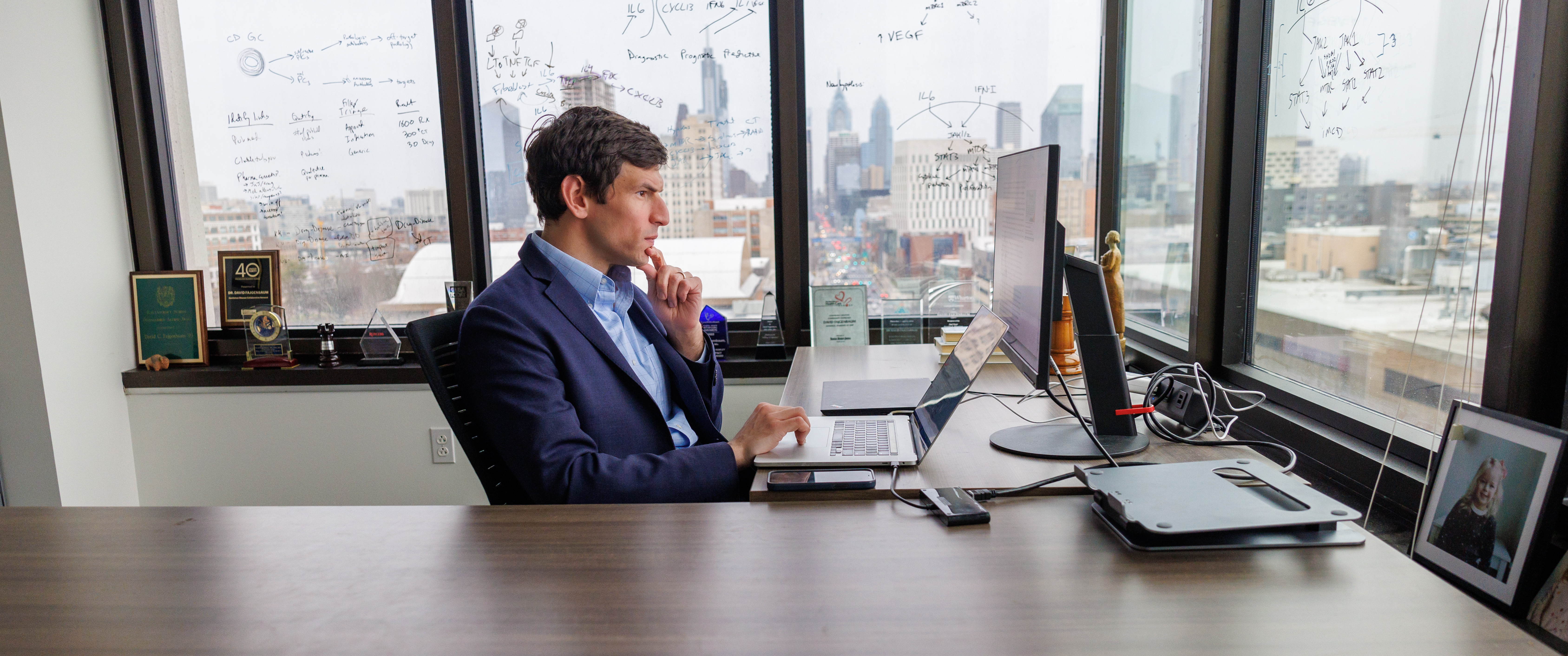 David Fajgenbaum, seated at a desk in profile, with his head pensively resting on his chin. Behind him, the windows are covered in equations and formulas written in dry-erase marker